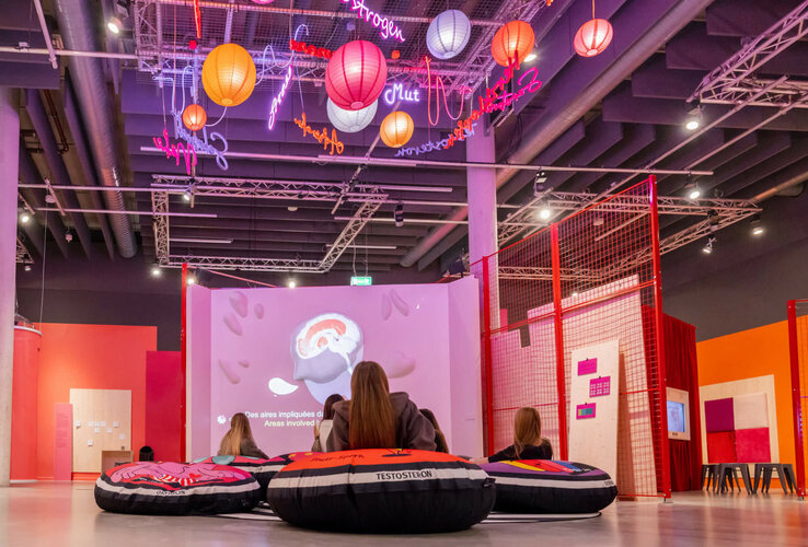 Besucher sitzen auf Sitzsäcken in einem modernen Raum mit bunten Lichtern und einer großen Leinwand.