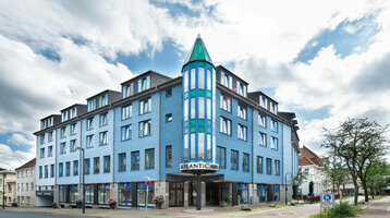 Blaues Gebäude des ATLANTIC Hotel Vegesack mit markantem Turm und Eingang, umgeben von Bäumen und Pflasterstraße.
