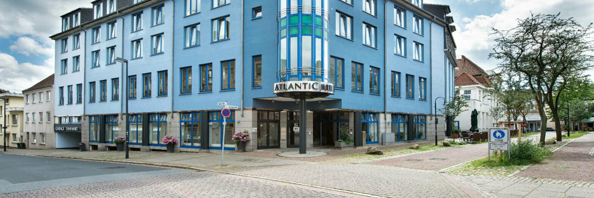 Blaues Gebäude des ATLANTIC Hotel Vegesack mit markantem Turm und Eingang, umgeben von Bäumen und Pflasterstraße.