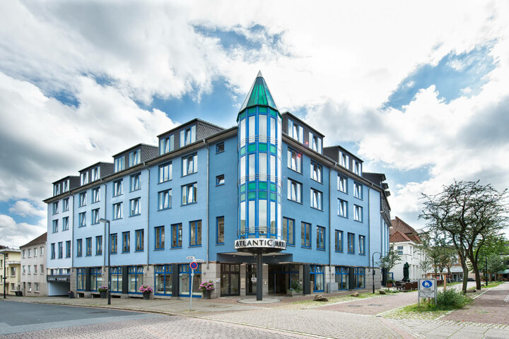 Blue building of the ATLANTIC Hotel Vegesack with striking tower and entrance, surrounded by trees and cobbled street.