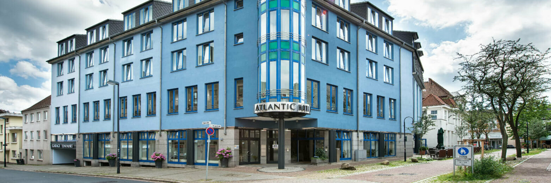 Blue building of the ATLANTIC Hotel Vegesack with striking tower and entrance, surrounded by trees and cobbled street.
