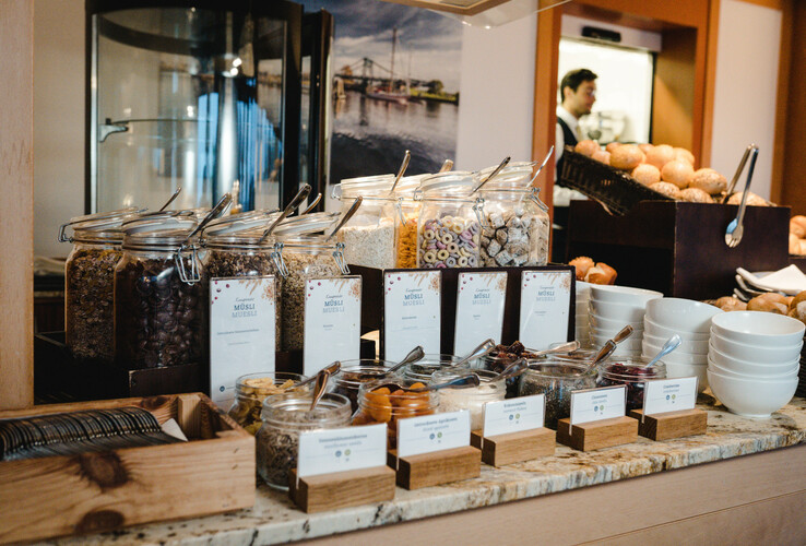 Breakfast buffet with cereal glasses, bowls, pastries and labels on a marble counter.
