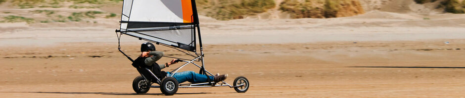 Person driving a beach sailing car on the beach, dunes in the background, blue sky.