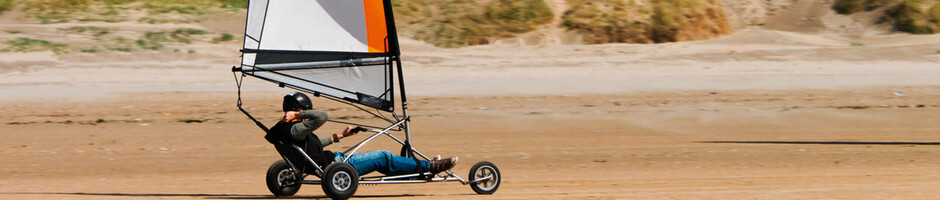 Person fährt Strandsegelwagen am Strand, Dünen im Hintergrund, blauer Himmel.