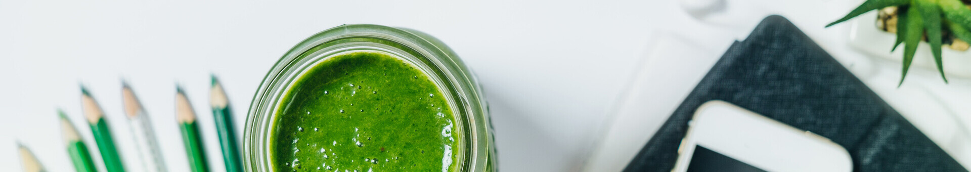 Grüner Smoothie im Glas, umgeben von grünen Buntstiften, einem Smartphone und einer kleinen Pflanze.