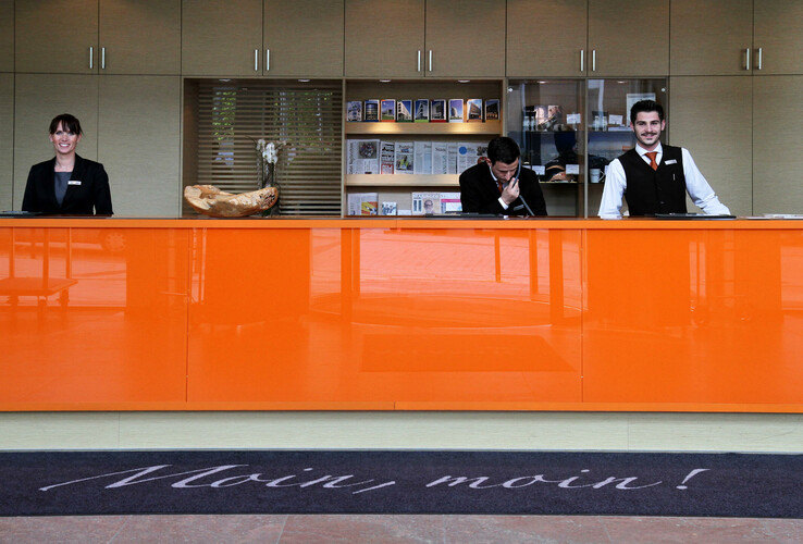 Rezeption im ATLANTIC Hotel Kiel mit drei lächelnden Mitarbeitern hinter einem orangefarbenen Tresen.