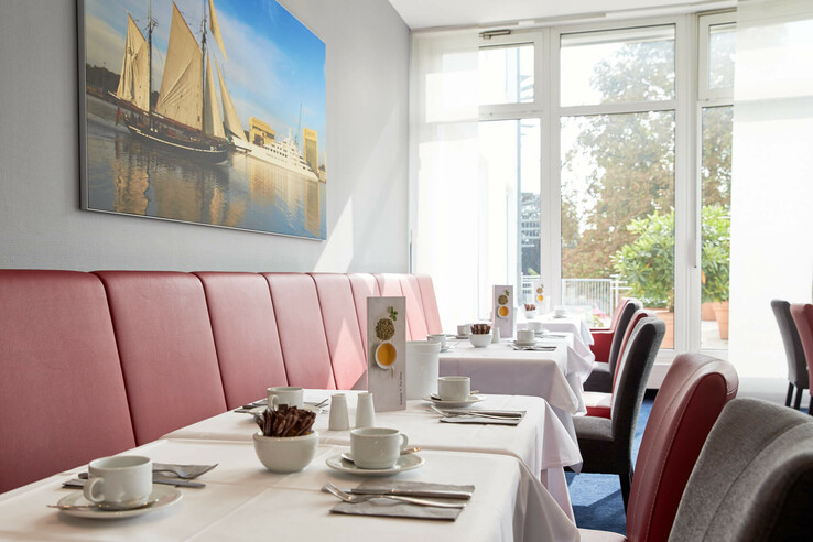 Bright breakfast room with red benches, laid tables and a large window in the ATLANTIC Hotel Vegesack.