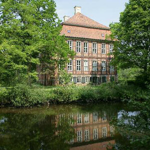Historic brick building on the waterfront, surrounded by green trees, reflected in the calm river.