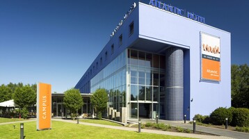ATLANTIC Hotel Universum in Bremen  Modernes Hotelgebäude mit blauer Fassade, großen Fenstern und gepflegtem Garten bei sonnigem Wetter.
