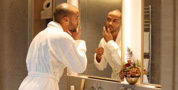 Mann im Bademantel pflegt sich im modernen Hotelbad mit Spiegel und Blumenarrangement.