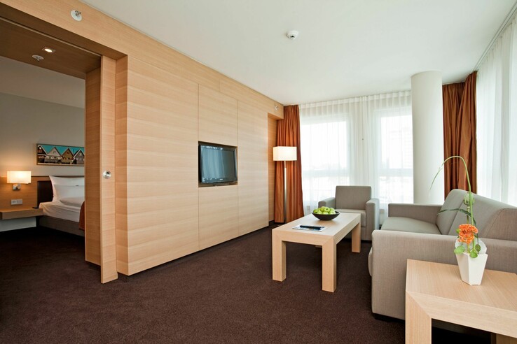 Modernes Hotelzimmer mit Sitzecke, grauem Sofa, Couchtisch, Flachbild-TV, hellem Holzdekor und großen Fenstern.