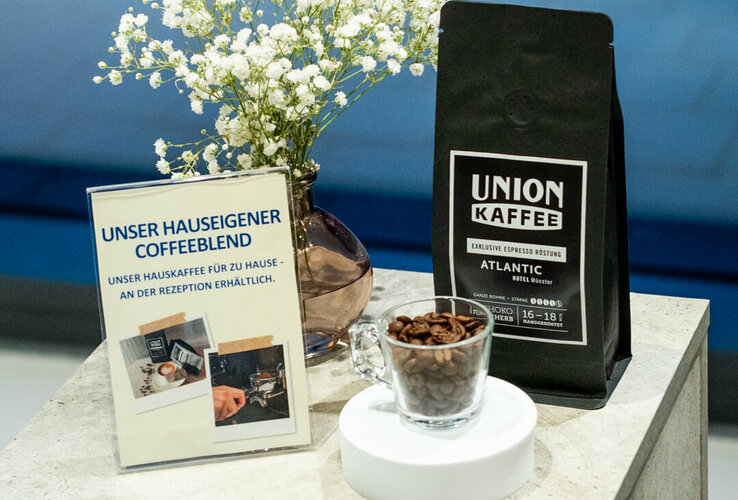 Ein Strauß Schleierkraut und eine kleine Espressotasse aus Glas mit Kaffeebohnen, daneben eine Packung Union Kaffee exklusive Espresso Röstung und ein Schild mit Text: "Unser hauseigener Coffeeblend. Unser Hauskaffee für zu Hause - an der Rezeption erhältlich.".
