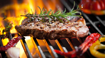 Grilled steak with rosemary on an open fire, surrounded by peppers and garlic on a grill rack.