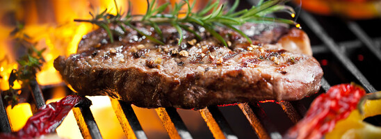 Grilled steak with rosemary on an open fire, surrounded by peppers and garlic on a grill rack.