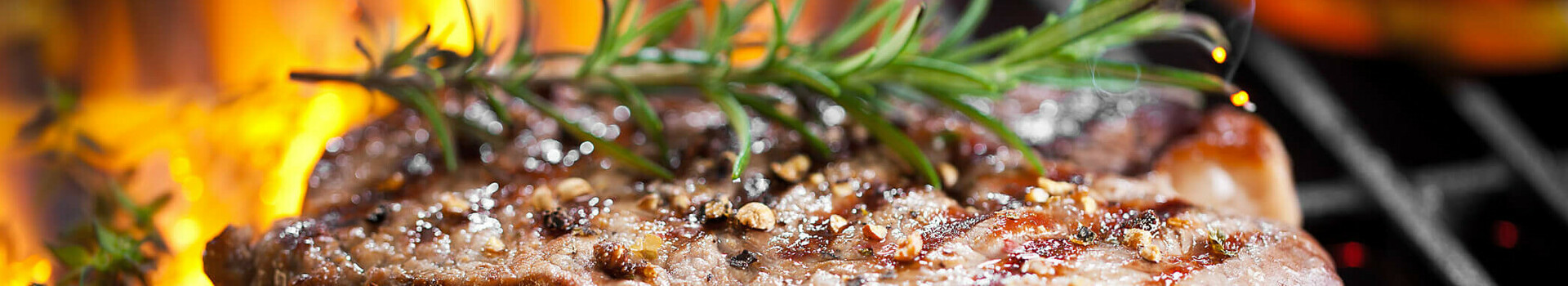 Gegrilltes Steak mit Rosmarin auf offenem Feuer, umgeben von Paprika und Knoblauch auf einem Grillrost.