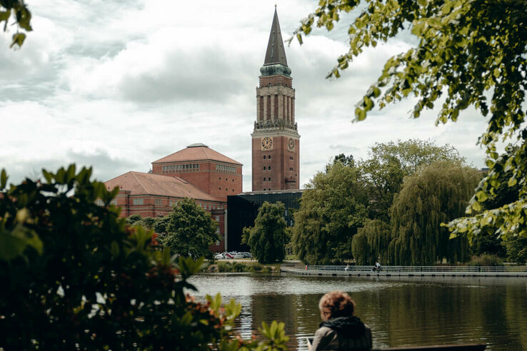 Frau sitzt auf Parkbank am See, im Hintergrund ein hoher Turm und Bäume.