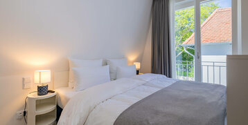 Bright hotel room with double bed, white bedding, bedside lamps and view of green trees through large windows.