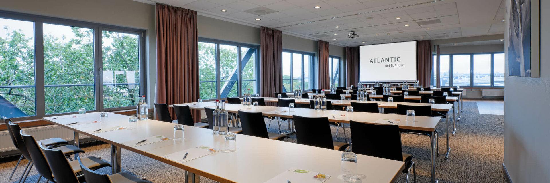 Modern conference room at the ATLANTIC Hotel Airport Bremen with tables, chairs and a large screen.