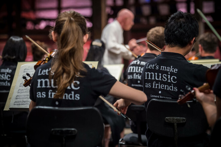 Ein Orchester spielt unter einem Dirigenten, Musiker tragen T-Shirts mit der Aufschrift "Let's make music as friends".
