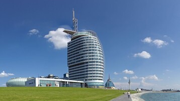 ATLANTIC Hotel Sail City in Bremerhaven Modernes Hotelgebäude am Wasser mit gläserner Fassade, umgeben von grünem Rasen und Promenade unter blauem Himmel.