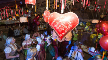 Festliche Hüttenszene mit tanzenden Menschen in Tracht, roten Herzdekorationen und gemütlicher Beleuchtung.