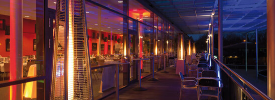 Beleuchtete Hotelterrasse bei Nacht mit Heizstrahlern, Tischen und Stühlen, Blick ins beleuchtete Restaurant.