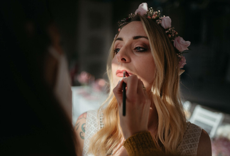 Eine Frau mit Blumenkranz wird in sanftem Licht geschminkt.