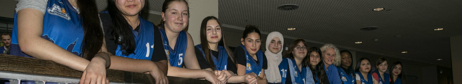 Kooperation Eisbären Bremerhaven U14 mit ATLANTIC Hotel Sail City Eine Gruppe junger Frauen in blauen Trikots lehnt an einem Geländer im ATLANTIC Hotel Sail City.