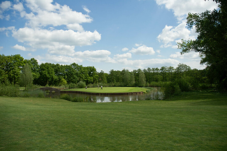 Grüne Golfanlage mit Teich und Brücke, umgeben von Bäumen, unter blauem Himmel mit Wolken.