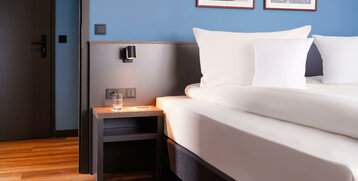 Modernes Hotelzimmer mit Doppelbett, blauen Wänden, Nachttisch, Glas Wasser und zwei gerahmten Bildern.