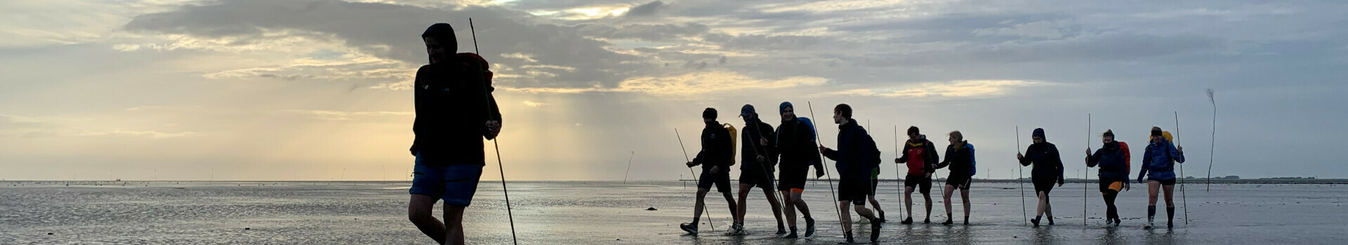 Gruppe von Menschen wandert bei Sonnenuntergang über das Watt, mit Wanderstöcken und Rucksäcken.