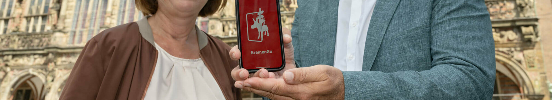Two people in front of a historic building hold a smartphone with the "BremenGo" app up to the camera.