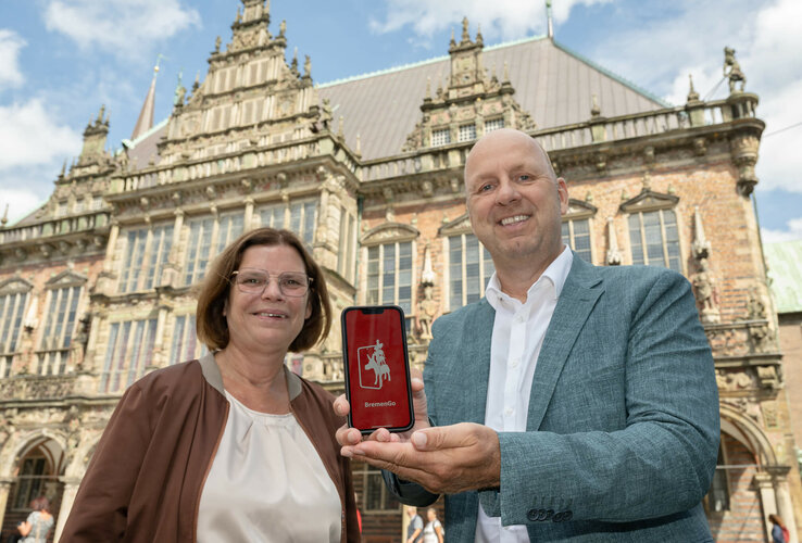 Two people in front of a historic building hold a smartphone with the "BremenGo" app up to the camera.