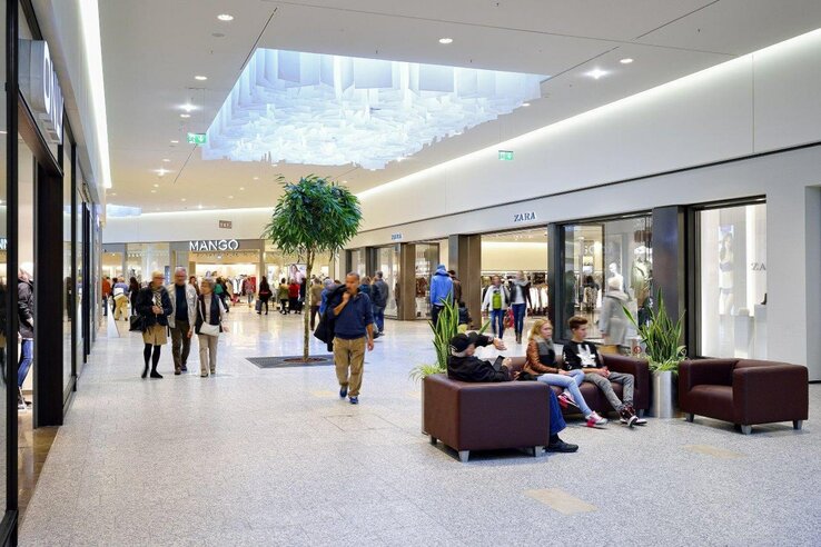 Einkaufszentrum mit Geschäften, Sitzbereich und Pflanzen; Menschen flanieren und entspannen auf Sofas.