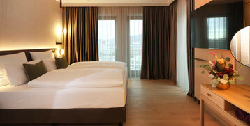 Modernes Hotelzimmer mit Doppelbett, stilvoller Beleuchtung, großem TV und Balkon mit Vorhängen im ATLANTIC Hotel Heidelberg.