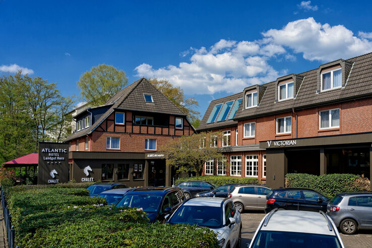 Brick hotel building with pitched roof, parking lot and green surroundings under a blue sky.