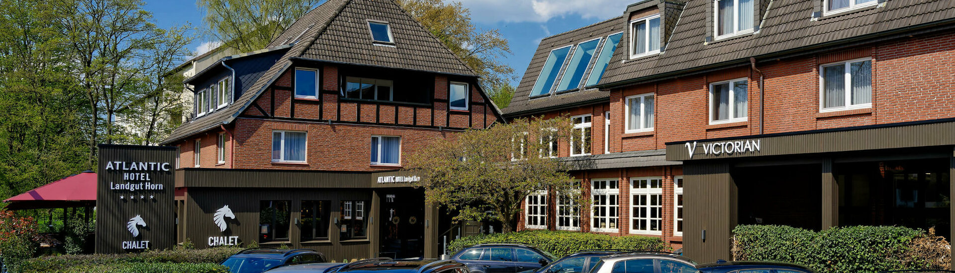 Außenansicht ATLANTIC Hotel Landgut Horn Bremen Backstein-Hotelgebäude mit Schrägdach, Parkplatz und grüner Umgebung unter blauem Himmel.