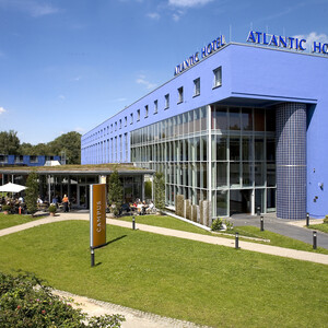 ATLANTIC Hotel Universum in Bremen: Modernes, blaues Gebäude mit Glasfassade, umgeben von Grünflächen und Außensitzbereich.