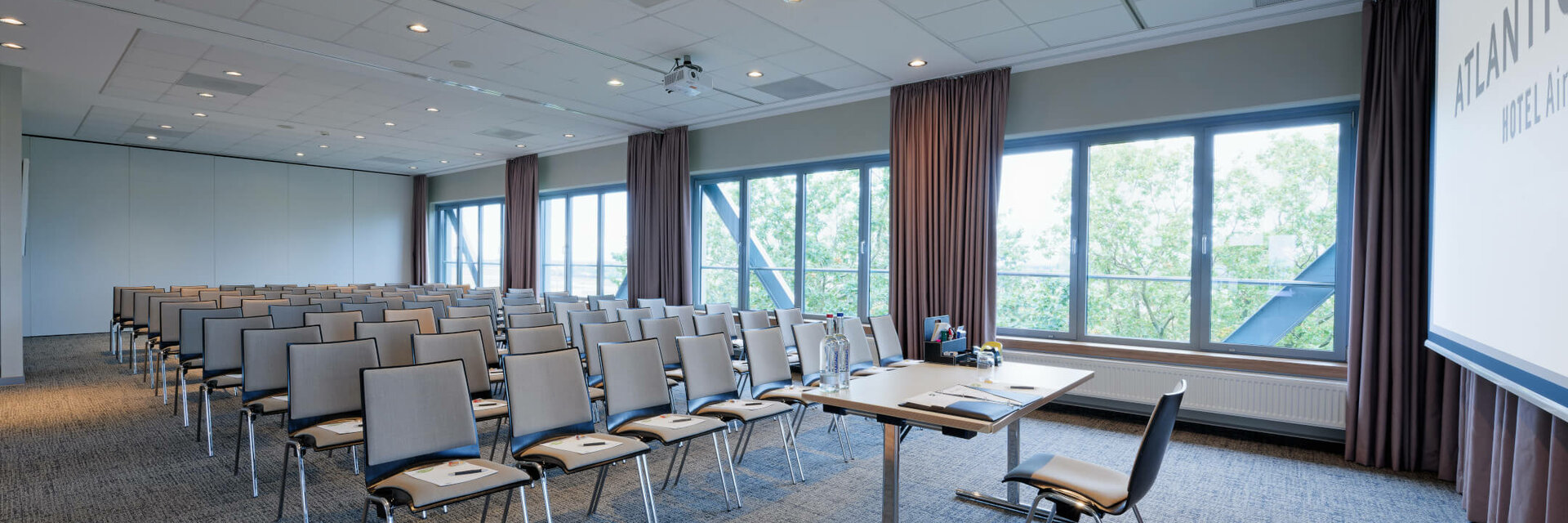 Heller Konferenzraum im ATLANTIC Hotel Airport Bremen mit Stuhlreihen, Tisch und großen Fenstern mit Blick ins Grüne.