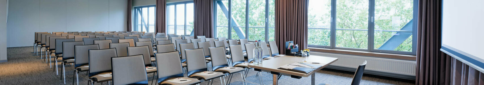 Heller Konferenzraum im ATLANTIC Hotel Airport Bremen mit Stuhlreihen, Tisch und großen Fenstern mit Blick ins Grüne.