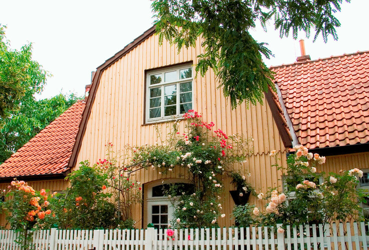 Gemütliches Haus mit Holzwänden, roten Ziegeldach, umgeben von blühenden Rosen und weißem Lattenzaun.