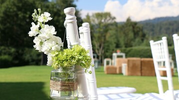 Weiße Gartenstühle mit Blumen in Glasvasen, auf grüner Wiese mit Wald im Hintergrund.