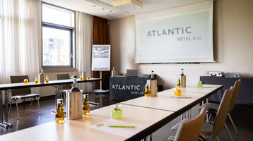 Veranstaltung Tagen im ATLANTIC Hotel Kiel Moderner Konferenzraum im ATLANTIC Hotel Kiel mit Tischen, Stühlen, Getränken und Leinwand.