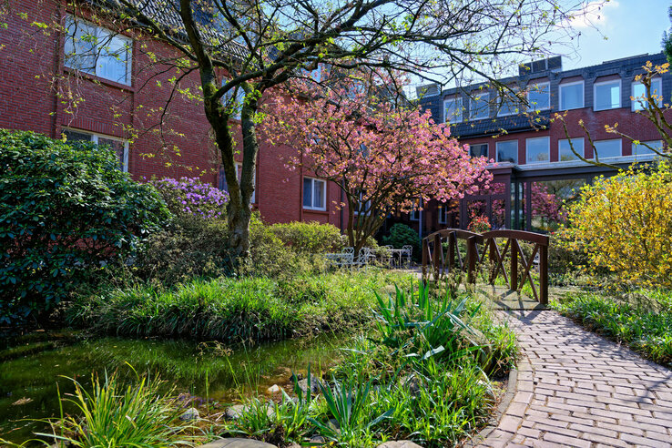 Gartenansicht mit blühenden Bäumen, kleinem Teich, Holzbrücke und rotem Backsteingebäude im ATLANTIC Hotel Landgut Horn.
