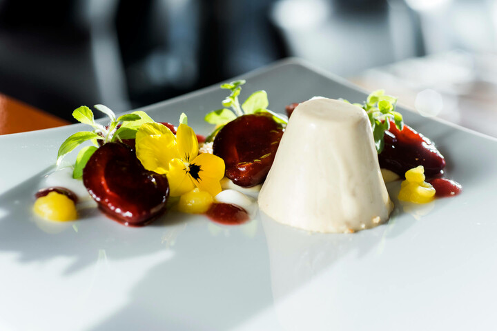 Speisen und Getränke | ATLANTIC Hotel Universum Bremen Elegantes Dessert mit Panna Cotta, roten Früchten und gelber Blüte auf weißem Teller, im ATLANTIC Hotel Universum.