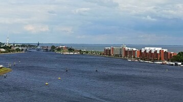Fluss mit Segelbooten, umgeben von Backsteingebäuden und grünem Ufer; bewölkter Himmel im Hintergrund.