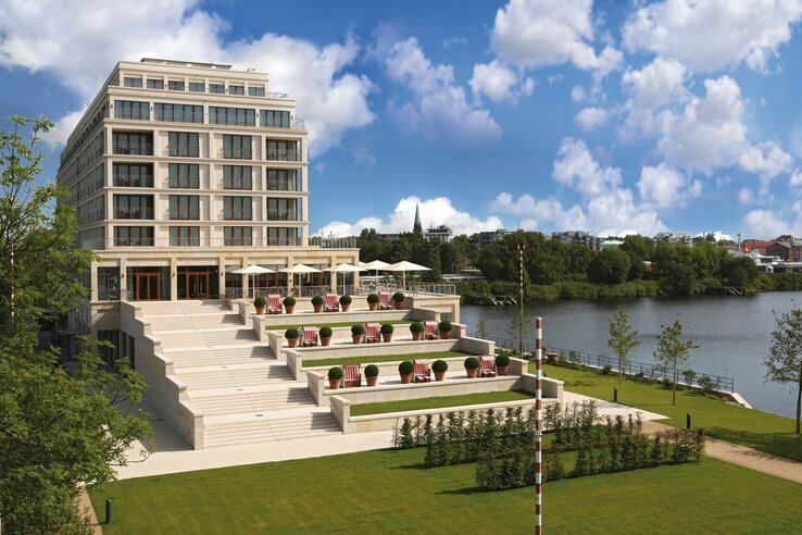 Modern hotel by the river with tiered terraces, sun loungers and parasols, surrounded by green countryside.