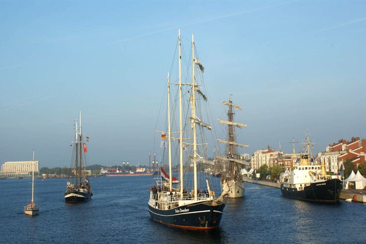Sailing ships in the harbor, surrounded by buildings and clear skies, with calm water.