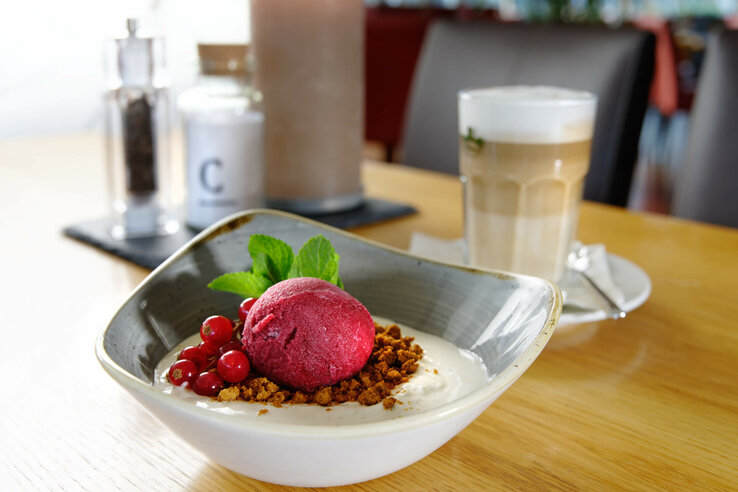 Rote Sorbetkugel mit Beeren und Minze auf Dessert, daneben Latte Macchiato auf Holztisch im ATLANTIC Hotel Universum.