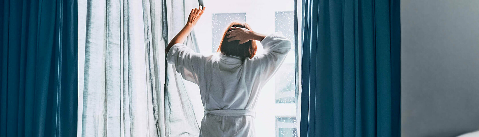 ATLANTIC Hotels – Aufstehen und wohlfühlen Person im Bademantel öffnet Vorhänge in einem Hotelzimmer, Tageslicht strömt herein, Laptop auf ungemachtem Bett.